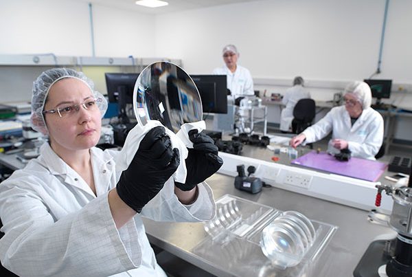 Woman in white coating in clean room holding up a large lens component