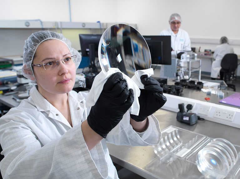 Quality-inspection-optics-768×572 woman wearing glassing in white coat holding up and inspecting large optical glass